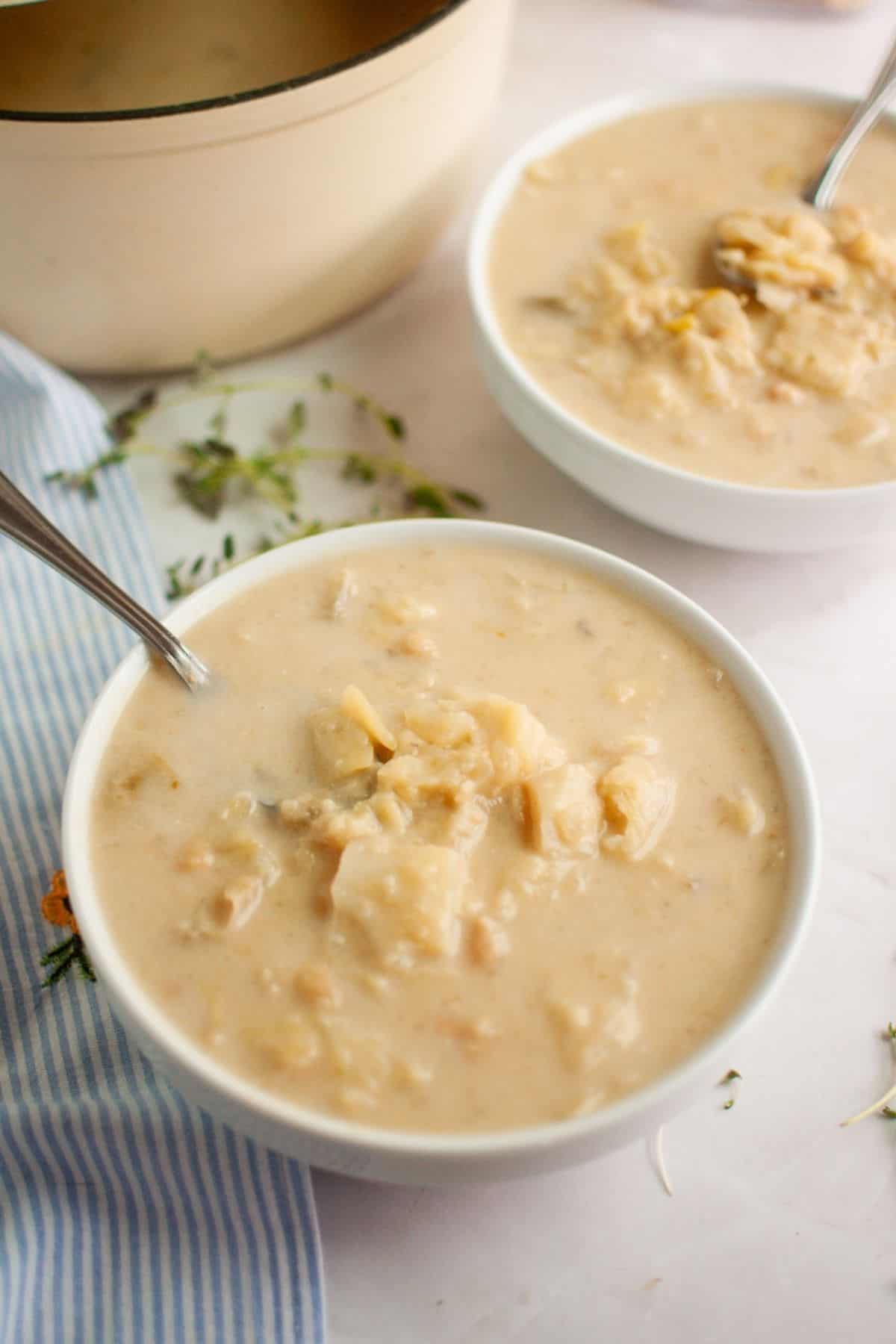 Two bowls of Cabbage and Bean Soup with chunks and spoons, placed on a white surface near a striped cloth and sprigs of thyme.