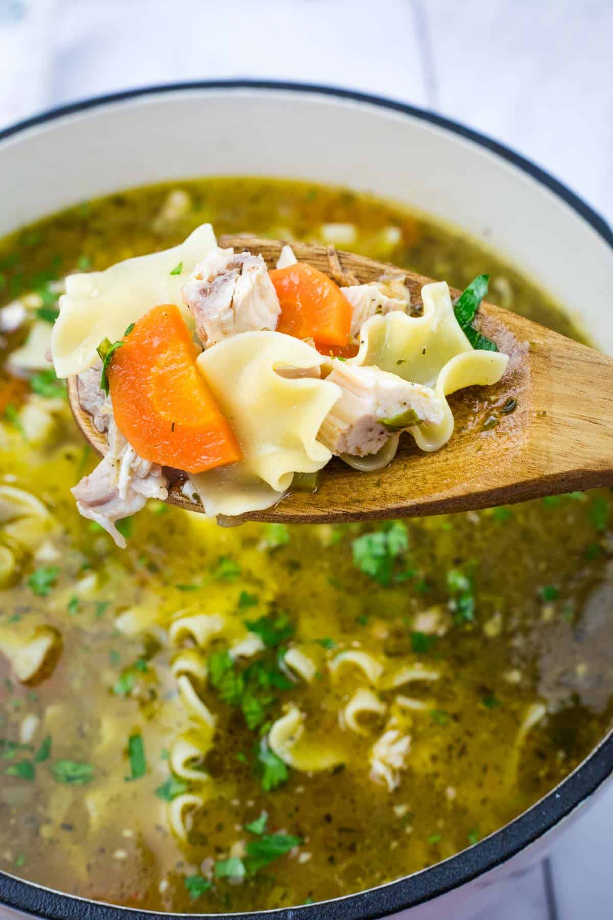 Wooden spoon with Rotisserie chicken noodle soup, featuring noodles, chicken pieces, and carrots, over a pot filled with broth and garnished with herbs.