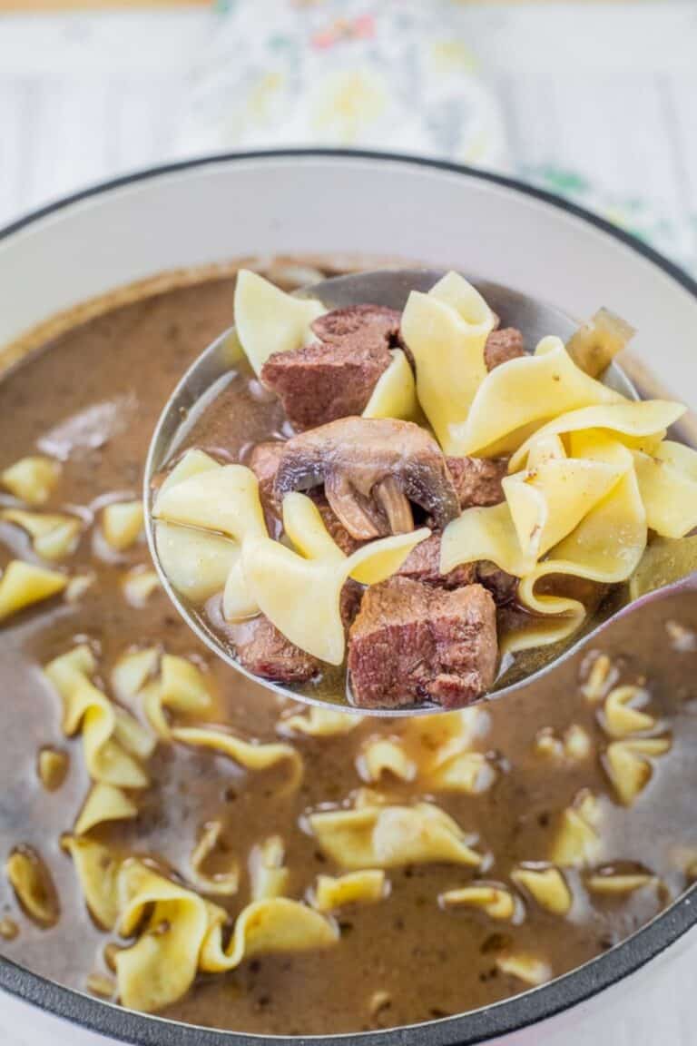 A ladle holding beef stew with noodles and mushrooms above a pot of the same stew.