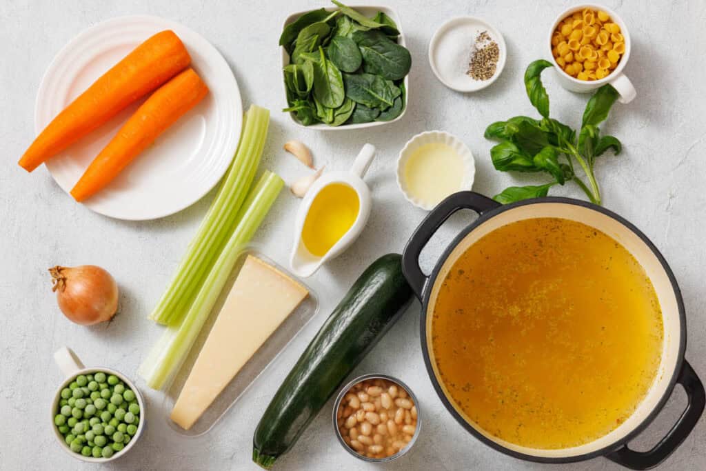 Ingredients for soup: carrots, spinach, pasta, salt, pepper, olive oil, celery, onion, Parmesan, zucchini, chickpeas, peas, and broth in a pot on a light surface.