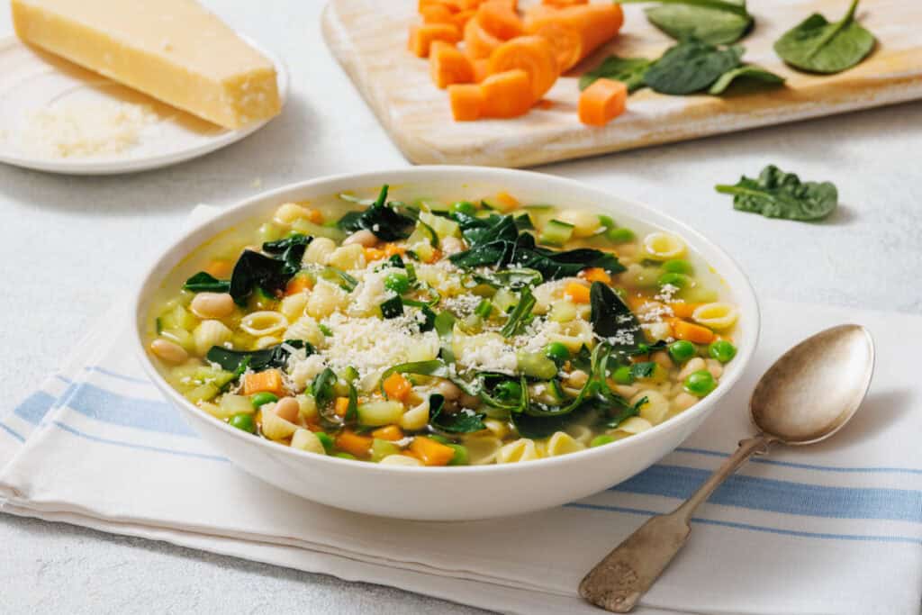 A bowl of Spring Minestrone with pasta shells, spinach, beans, and peas, topped with grated cheese. Fresh carrots and spinach on a cutting board nearby. Spoon and napkin beside the bowl.