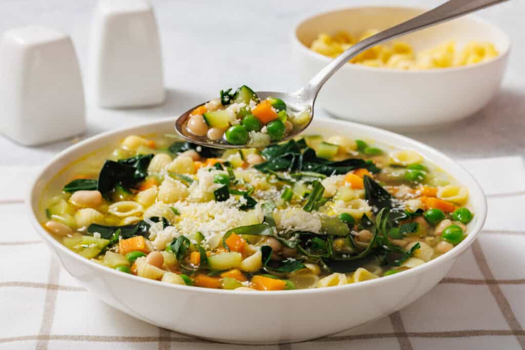 A bowl of Spring Minestrone with pasta, peas, carrots, and greens on a checkered cloth. A spoon holds a portion above it. Two salt shakers and a bowl are in the background.