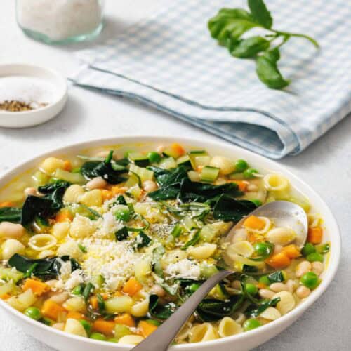 A bowl of Spring Minestrone with shell pasta, carrots, spinach, peas, and topped with grated cheese. A spoon rests in the bowl. A jar of salt and a checked napkin are in the background.