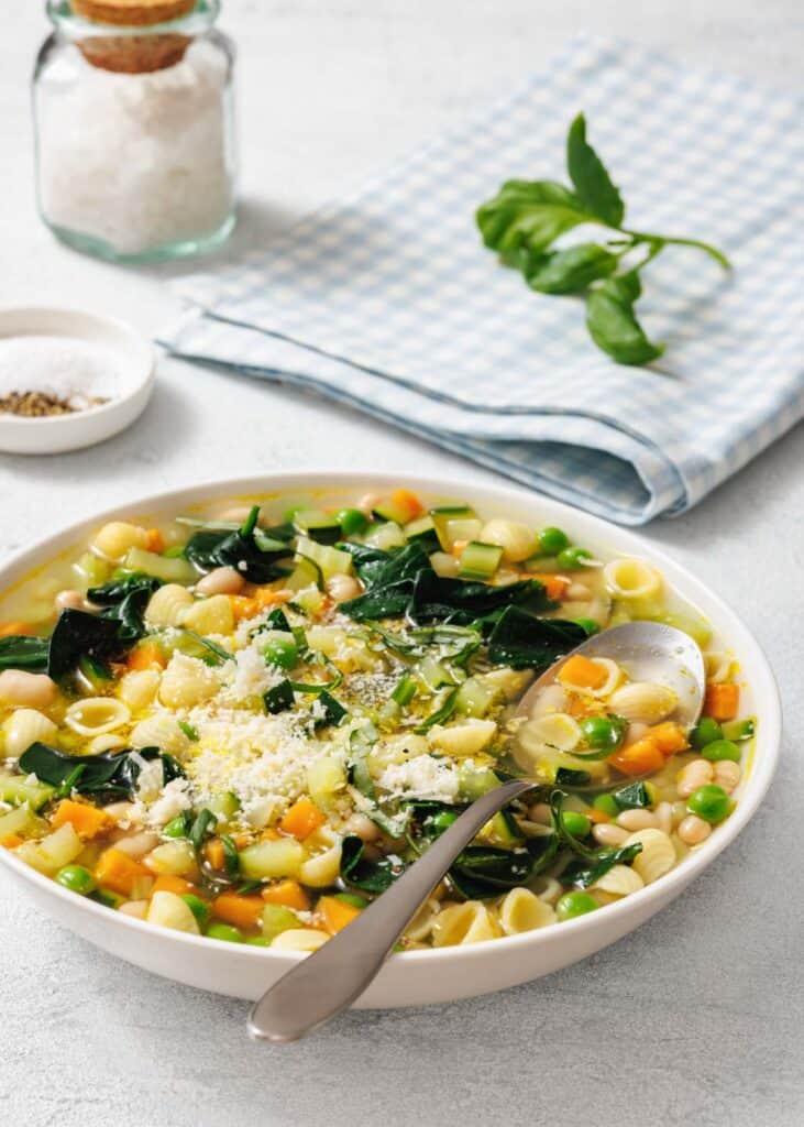 A bowl of Spring Minestrone with shell pasta, carrots, spinach, peas, and topped with grated cheese. A spoon rests in the bowl. A jar of salt and a checked napkin are in the background.
