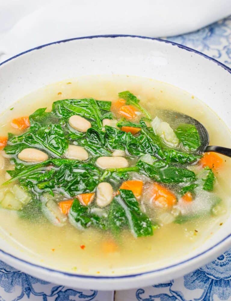 A bowl of Rice and White Bean Soup containing white beans, kale, carrots, and other vegetables, with a spoon on the side.