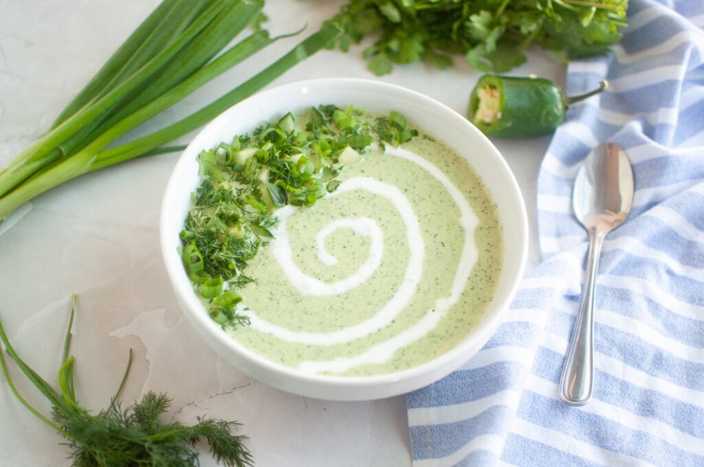Overhead shot of a bowl of creamy green soup with herbs and a swirl of cream, alongside a spoon, cloth, and vegetables.