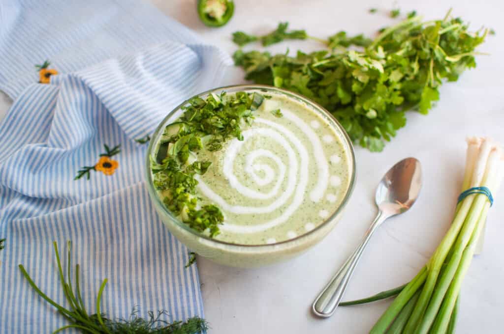 Overhead shot of a bowl of creamy green soup with herbs and a swirl of cream, alongside a spoon, cloth, and vegetables.