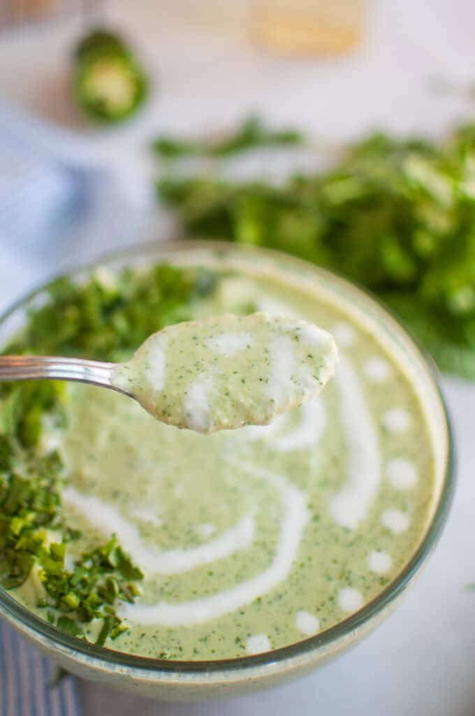 A spoonful of creamy green soup is lifted above a clear bowl filled with the same soup, garnished with herbs and a swirl of cream.