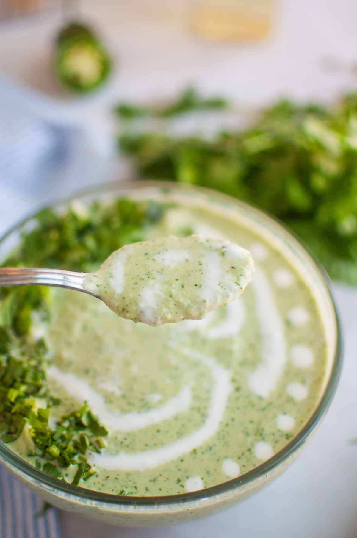 A spoonful of creamy green soup is lifted above a clear bowl filled with the same soup, garnished with herbs and a swirl of cream.