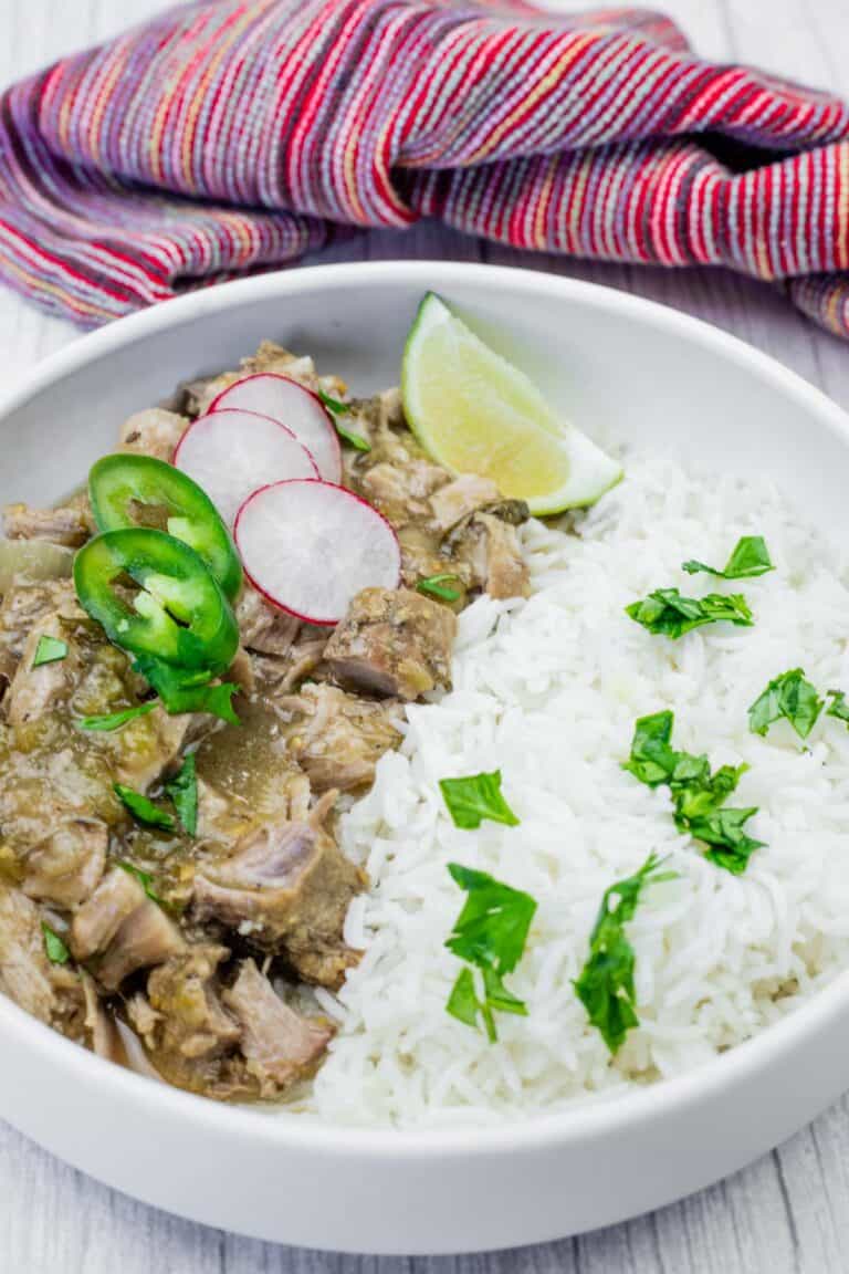 A bowl of white rice topped with chopped herbs, served with shredded meat in green sauce, garnished with radish slices, jalapeño, and a lime wedge.
