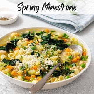 A bowl of Spring Minestrone with shell pasta, carrots, spinach, peas, and topped with grated cheese. A spoon rests in the bowl. A jar of salt and a checked napkin are in the background.