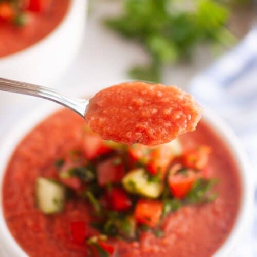 A spoonful of gazpacho held above a bowl of gazpacho garnished with diced vegetables and herbs.