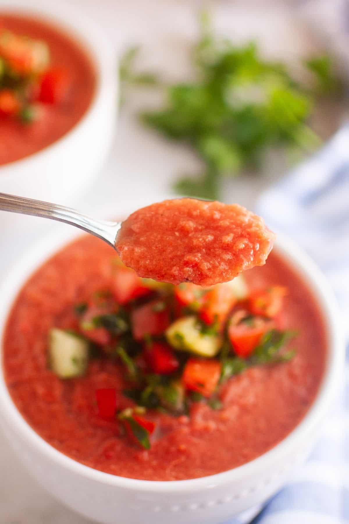 A spoonful of gazpacho held above a bowl of gazpacho garnished with diced vegetables and herbs.