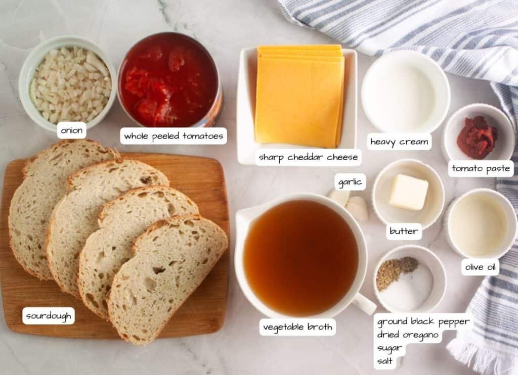 A top-down view of ingredients for a recipe, including sourdough bread, chopped onion, whole peeled tomatoes, cheddar cheese slices, heavy cream, tomato paste, garlic, butter, olive oil, broth, and seasonings.