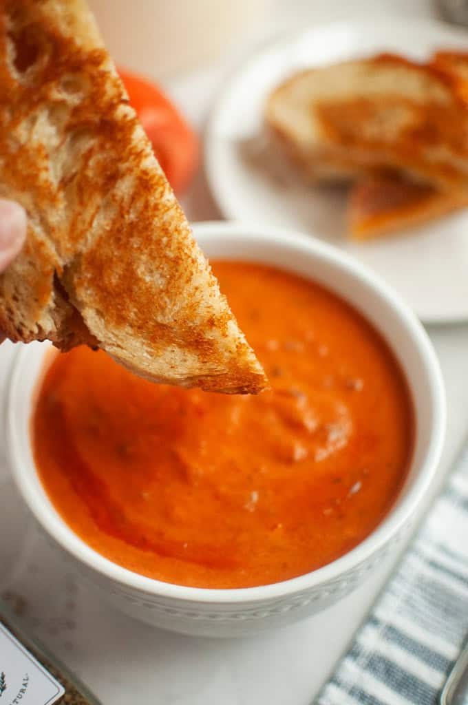 A hand dipping a piece of grilled cheese sandwich into a bowl of creamy tomato soup.