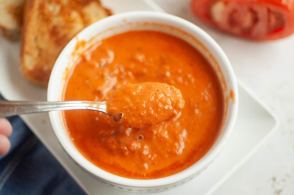 A spoonful of creamy orange-red tomato soup over a white bowl filled with more soup.