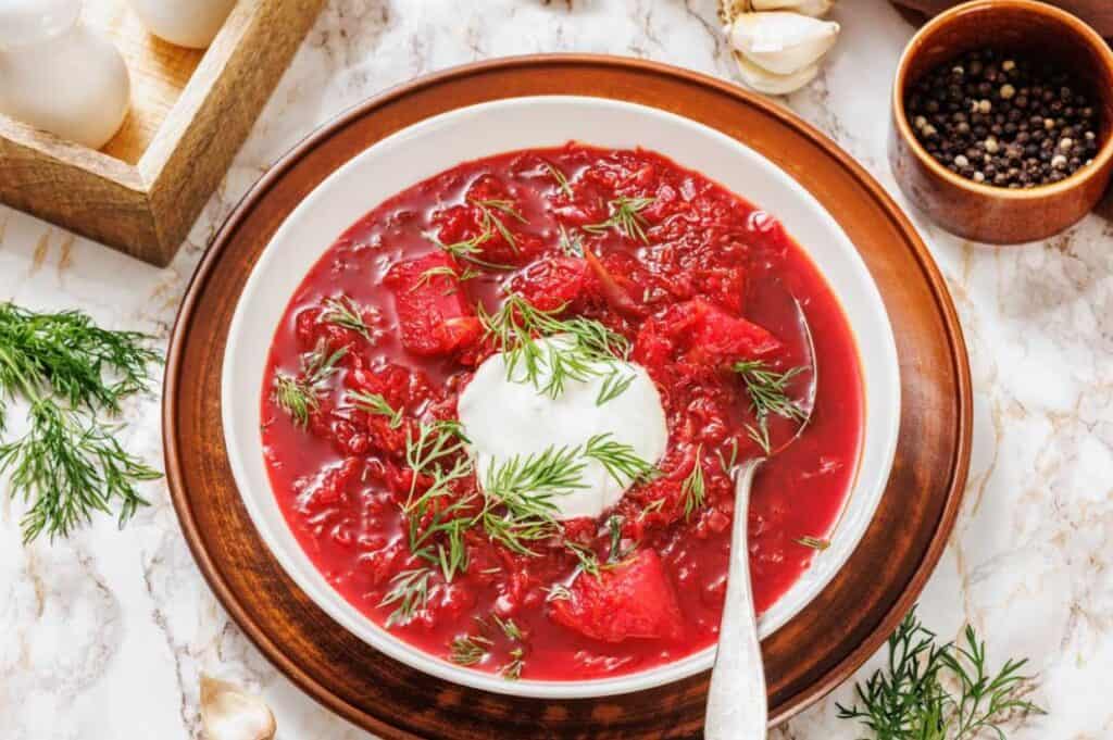 A bowl of beet soup garnished with fresh dill and a dollop of sour cream, served with a spoon on a wooden plate.