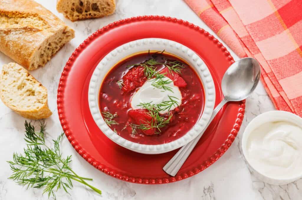 A bowl of borscht topped with sour cream and dill is served on a red plate with a spoon, bread slices, extra dill, and a small bowl of sour cream on the side.