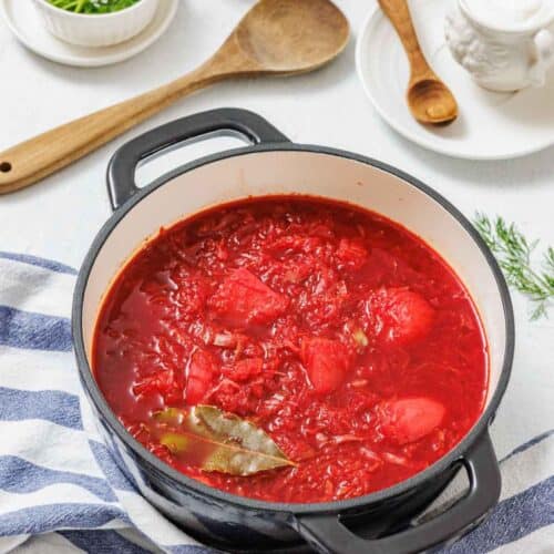 A black pot filled with red borscht soup sits on a striped cloth, surrounded by herbs, a wooden spoon, and small bowls with condiments.