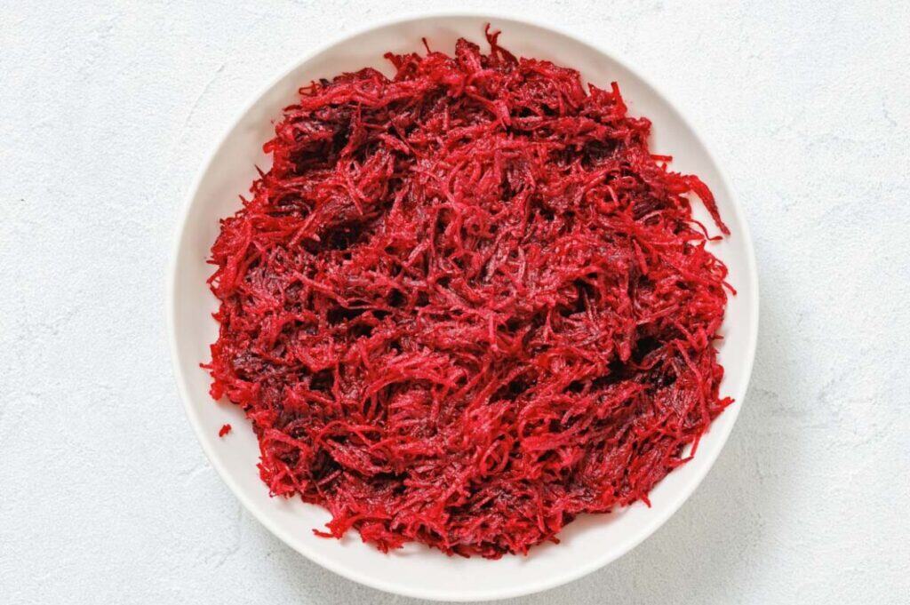 A white bowl filled with finely shredded raw beets, placed on a light-colored surface.