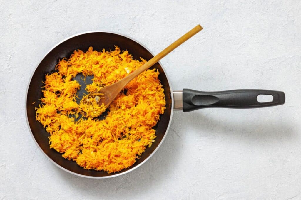 Shredded carrots being sautéed in a black frying pan with a wooden fork on a light surface.