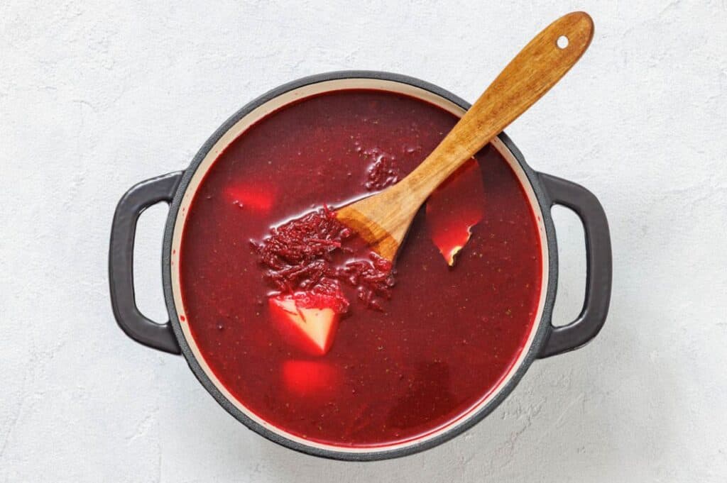 A pot of red beet soup with visible potato chunks, stirred with a wooden spoon, on a light surface.