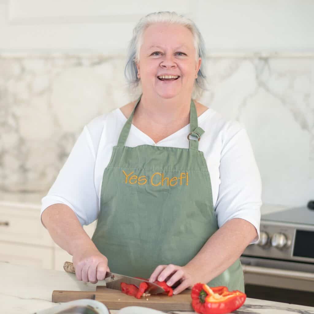 Chef Jenn in a green "Yes Chef!" apron slices a red bell pepper on a cutting board in a modern kitchen.