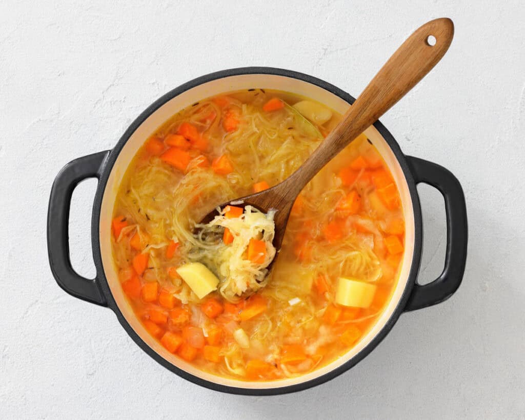 A pot of vegetable soup with carrots, potatoes, and noodles, being stirred with a wooden spoon on a light surface.