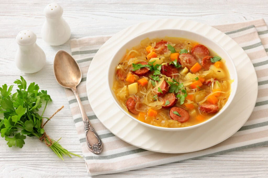 A bowl of vegetable and sausage soup garnished with parsley, placed on a striped cloth beside a spoon, fork, salt, pepper shakers, and a bunch of fresh parsley.