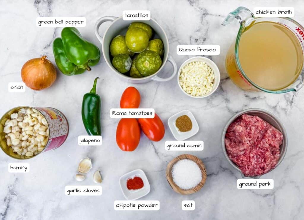 Ingredients for a recipe arranged on a white surface, including ground pork, hominy, vegetables, spices, queso fresco, tomatillos, and chicken broth, each labeled with text.