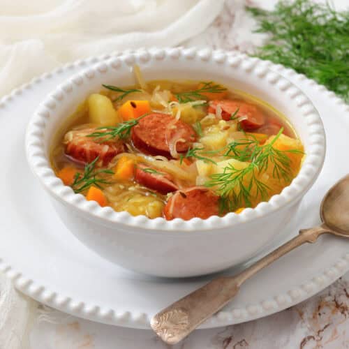 A white bowl of soup with sausage slices, potatoes, carrots, cabbage, and dill garnish, placed on a white plate with a spoon beside it. Fresh dill is in the background.