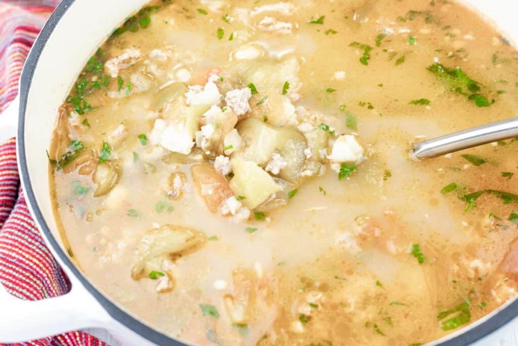 A pot of soup with ground meat, vegetables, herbs, and broth, with a metal ladle resting inside.