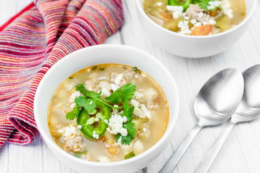 Two bowls of soup garnished with cilantro, sliced jalapeño, and cheese, placed on a white surface with two spoons and a striped napkin beside them.