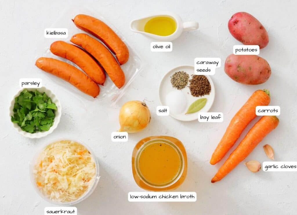 Ingredients for a dish, including kielbasa, olive oil, potatoes, carrots, garlic, onion, parsley, sauerkraut, chicken broth, caraway seeds, bay leaf, and salt, arranged on a white surface.
