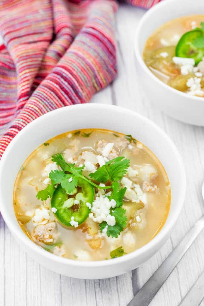 A bowl of soup with cilantro, sliced jalapeño, and crumbled cheese on top, next to a colorful cloth napkin and a spoon on a white wooden surface.