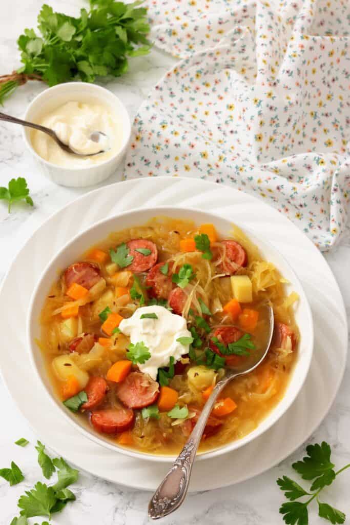 A bowl of vegetable and sausage soup garnished with parsley and a dollop of sour cream, with a spoon inside, next to a small bowl of sour cream and fresh parsley on a white floral napkin.