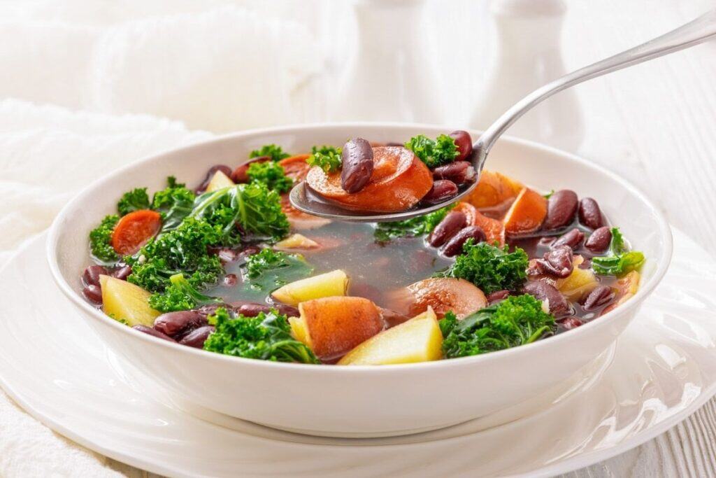 A bowl of vegetable soup with potatoes, kielbasa, kale, and kidney beans, with a spoon lifting a portion from the bowl.