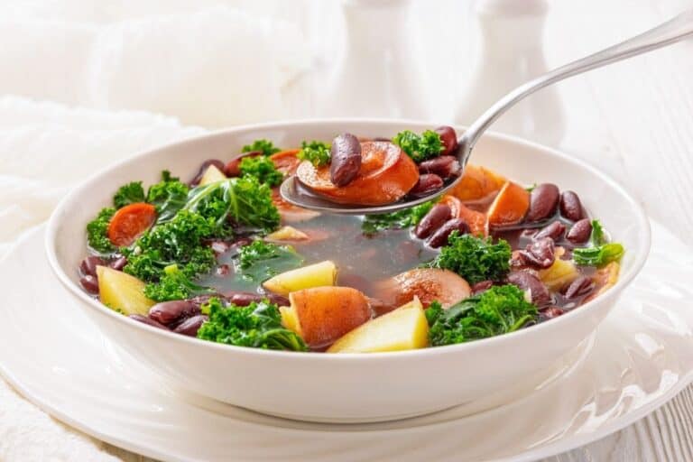 A bowl of vegetable soup with potatoes, kielbasa, kale, and kidney beans, with a spoon lifting a portion from the bowl.