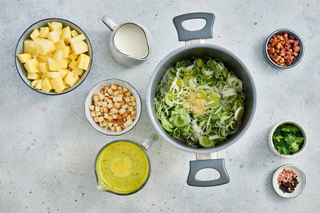 Top-down view of ingredients for leek and potato soup, including chopped potatoes, sliced leeks in a pot, cream, croutons, bacon, herbs, and seasonings arranged on a gray surface.