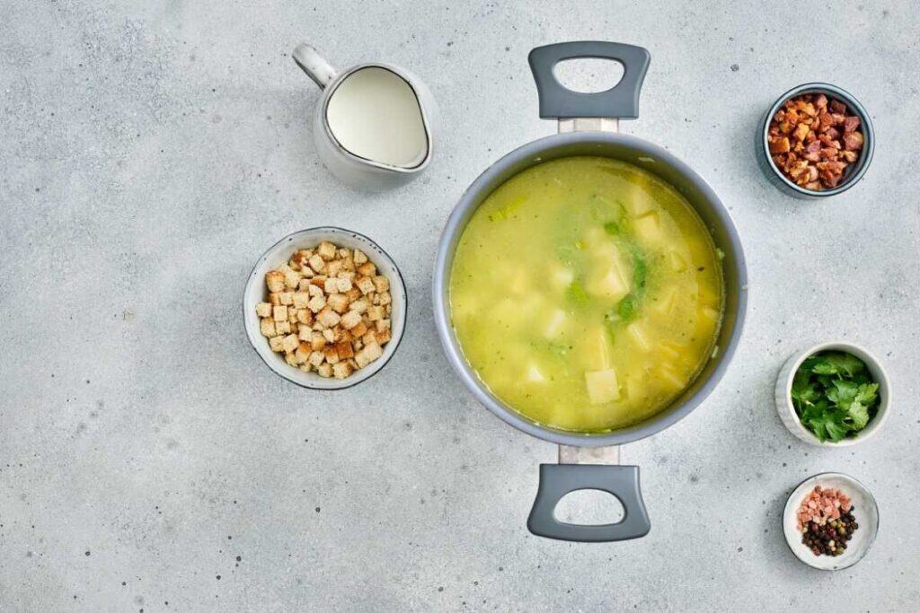 A pot of vegetable soup with potatoes sits on a countertop, surrounded by bowls of croutons, cream, herbs, bacon bits, and seasoning.