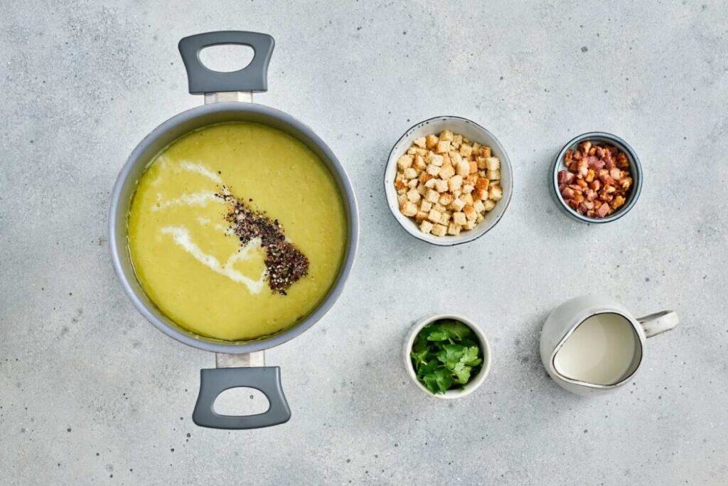 A pot of creamy soup topped with pepper and cream, next to bowls of croutons, bacon bits, parsley, and a jug of cream on a gray surface.