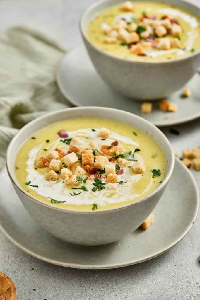 Two bowls of creamy soup topped with croutons, herbs, and bacon bits, served on matching saucers with a napkin beside them.