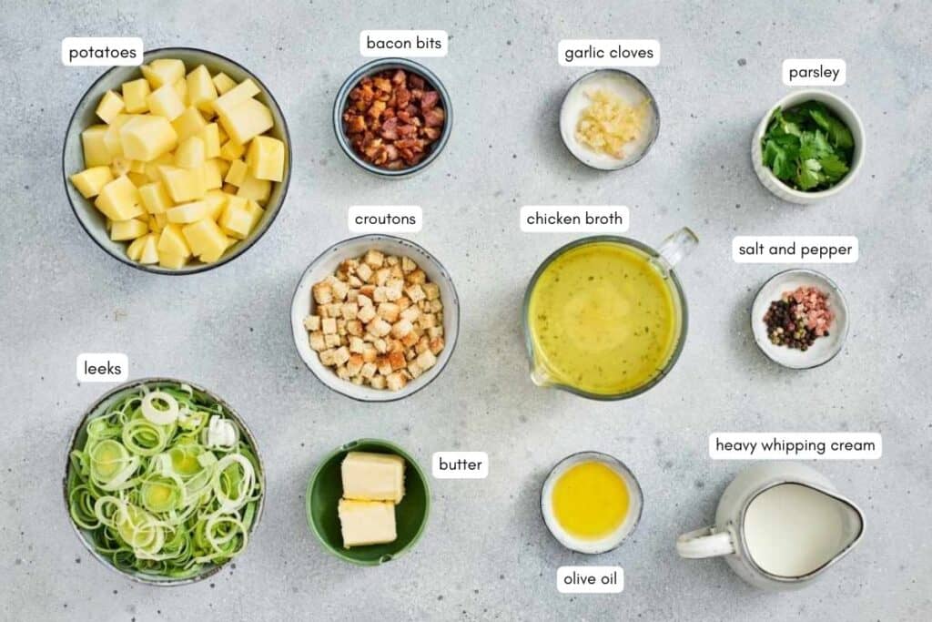 Top-down view of bowls containing potatoes, leeks, butter, croutons, bacon bits, garlic, chicken broth, olive oil, heavy cream, parsley, and salt and pepper.