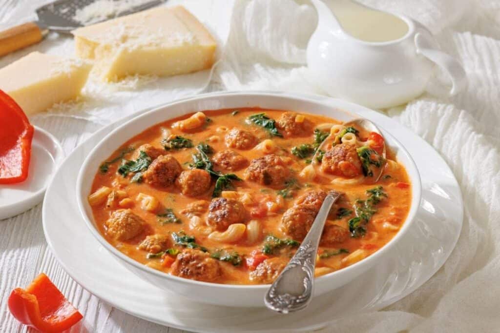 A bowl of meatball soup with pasta, spinach, and tomato broth sits on a table beside a spoon, cheese wedges, a grater, a creamer, and a piece of red bell pepper.