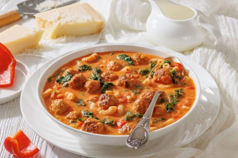 A bowl of meatball soup with pasta, spinach, and tomato broth sits on a table beside a spoon, cheese wedges, a grater, a creamer, and a piece of red bell pepper.