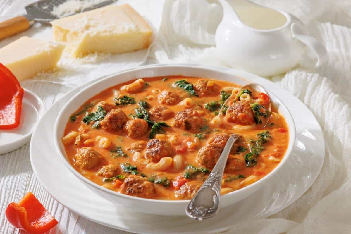 A bowl of meatball soup with pasta, spinach, and tomato broth sits on a table beside a spoon, cheese wedges, a grater, a creamer, and a piece of red bell pepper.