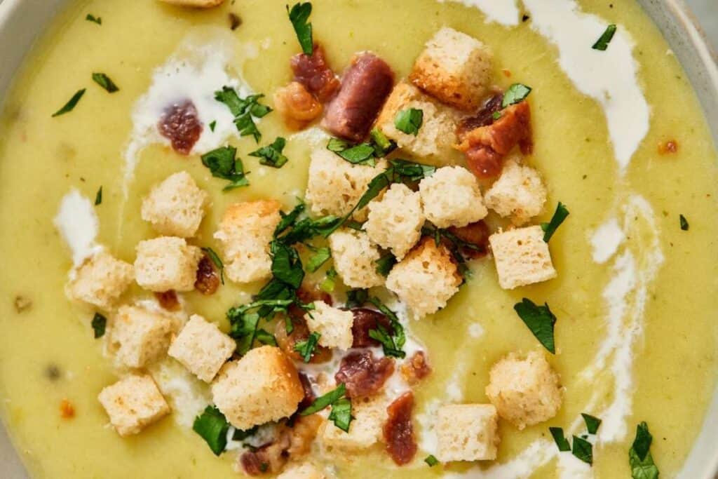 A close-up of creamy soup garnished with croutons, chopped herbs, pieces of bacon, and a swirl of cream.
