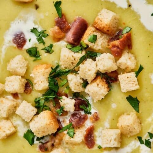 A close-up of creamy soup garnished with croutons, chopped herbs, pieces of bacon, and a swirl of cream.