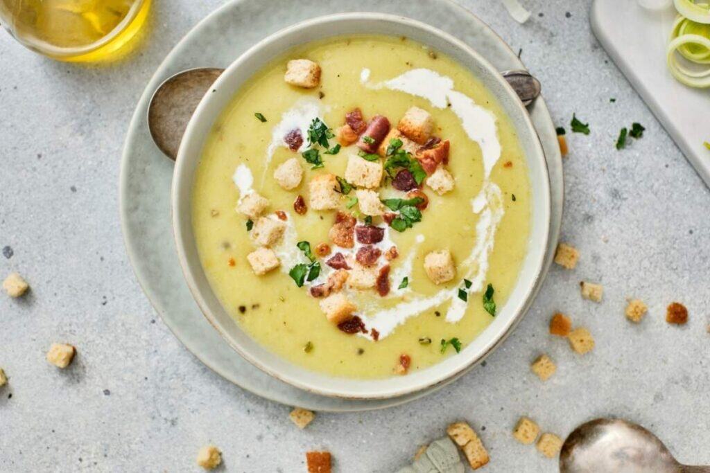A bowl of creamy soup garnished with croutons, bacon bits, chopped herbs, and a swirl of cream, placed on a plate with scattered croutons around.