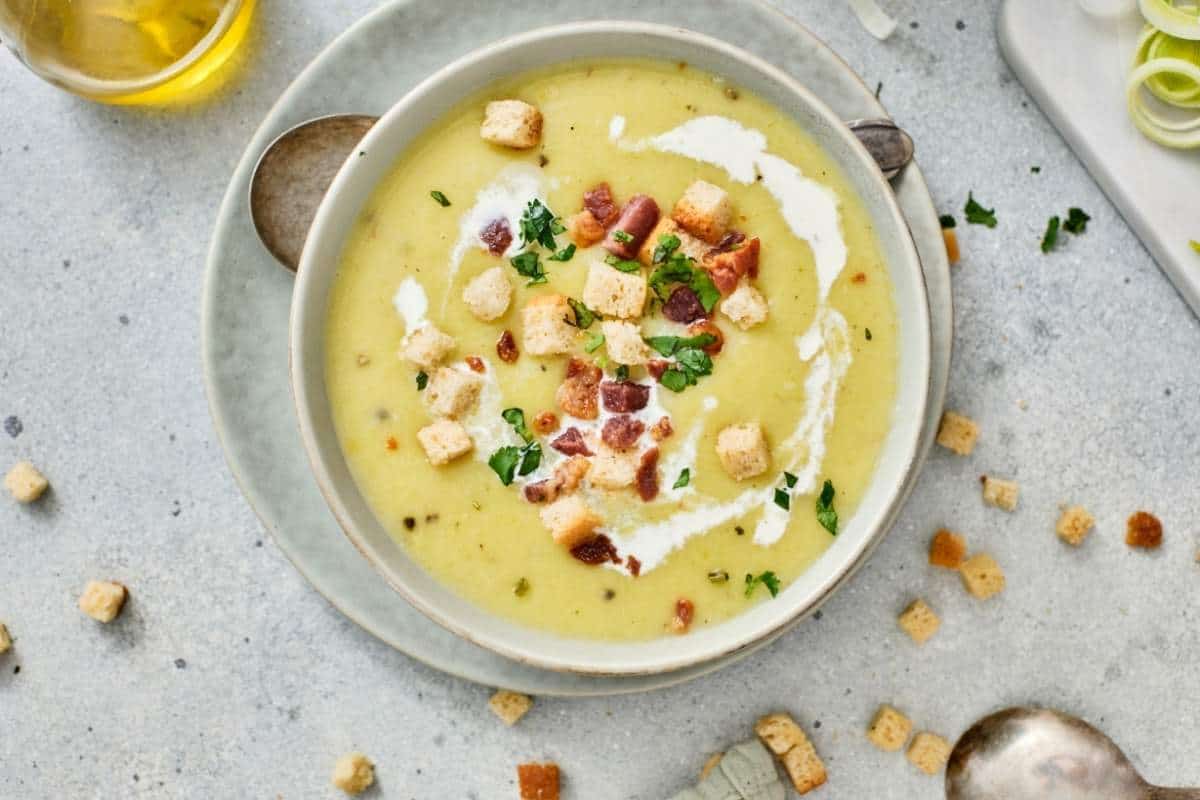 A bowl of creamy soup garnished with croutons, bacon bits, chopped herbs, and a swirl of cream, placed on a plate with scattered croutons around.